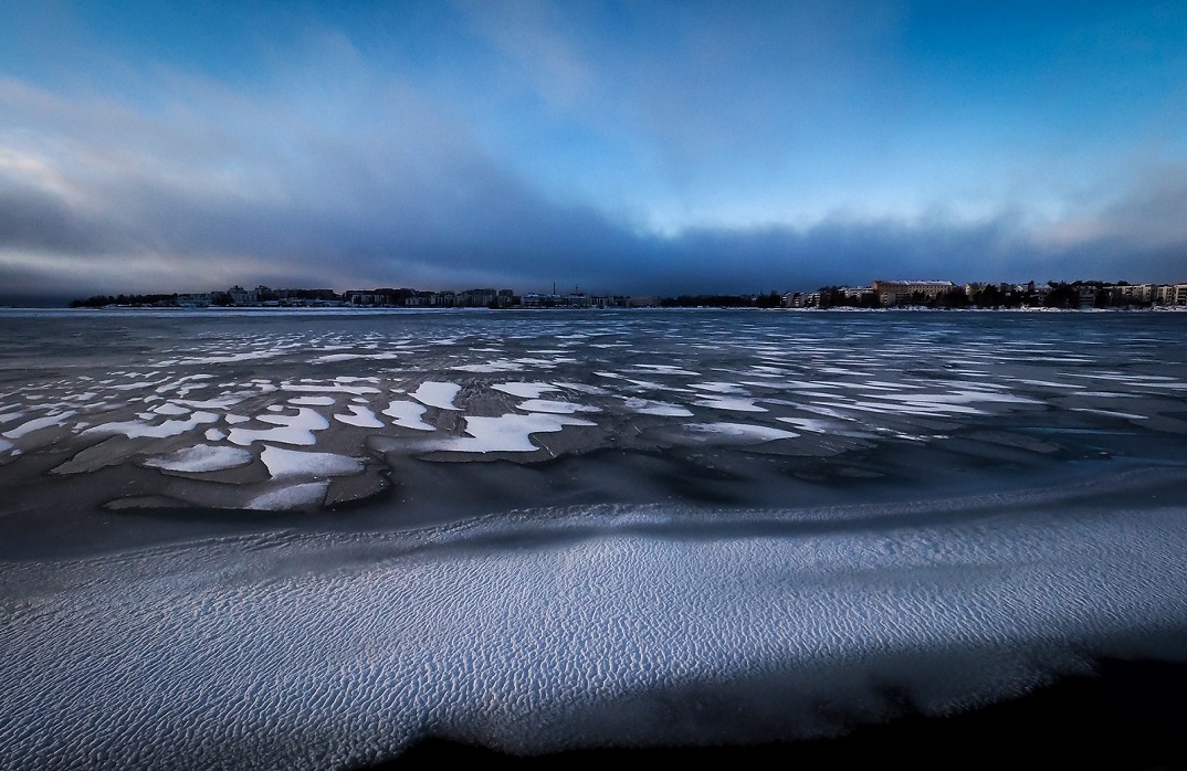 Maisemakuva järven rannalta, jossa vastarannalla kaupunki ja rannalla on hieman lunta ja jäätä.